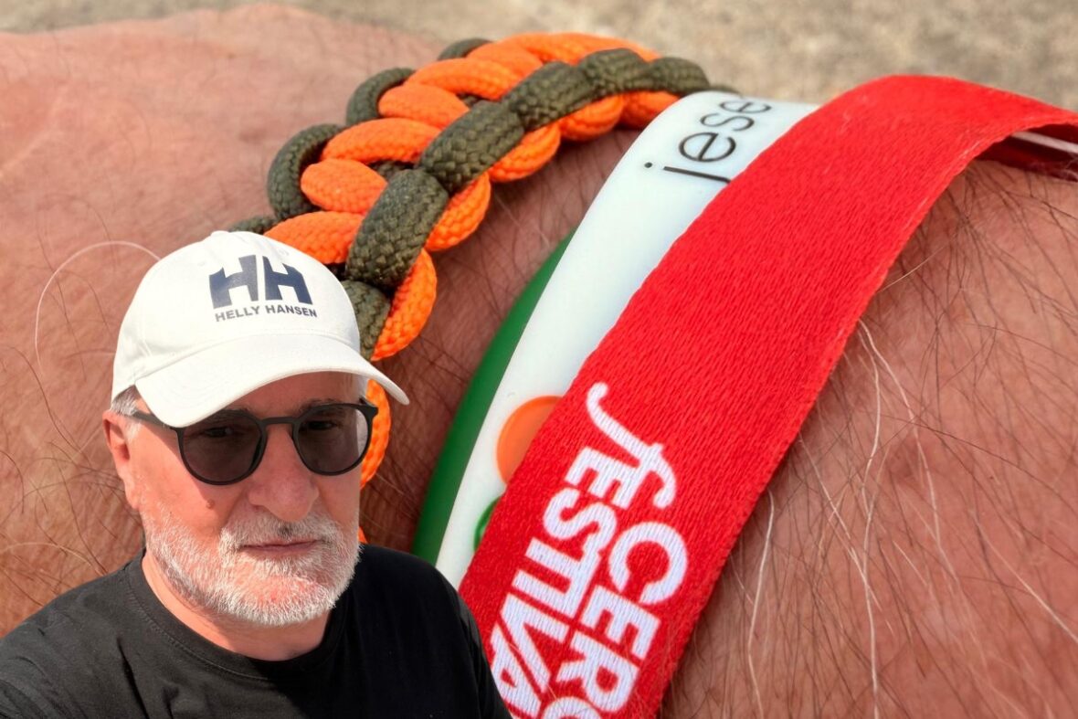 Bliskie ujęcie Starego Dziada w białej czapce Helly Hansen, czarnych okularach i czarnej koszulce, z plecioną bransoletką na przedramieniu na tle innych kolorowych opasek festiwalowych